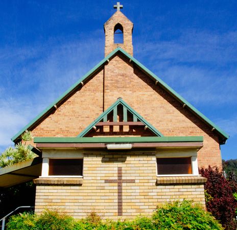 Front View Of A Brick Church With Green Roof And Cross Against A Clear Blue Sky