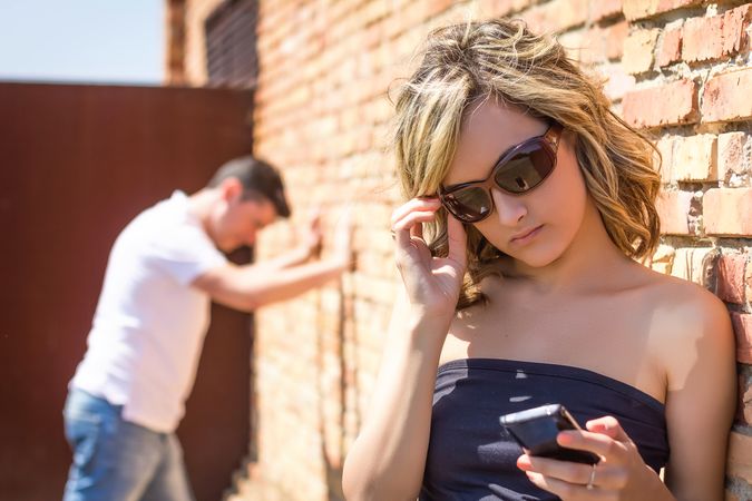 Angry woman looking phone and man posing on wall