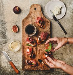 Hands holding canapé with prosciutto, goat cheese and grilled figs over wooden board