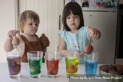 Girl and toddler putting eggs in coloring glasses in the kitchen at home 0KQ8V4