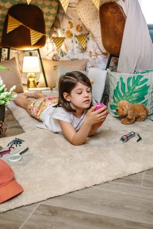 Little girl using walkie-talkie lying over rug surrounded by cushions and plants in teepee at home