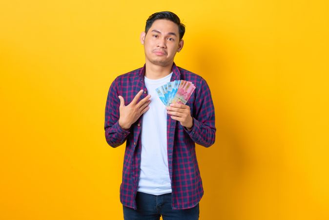 Sad Asian man in plaid shirt  pointing while holding banknotes