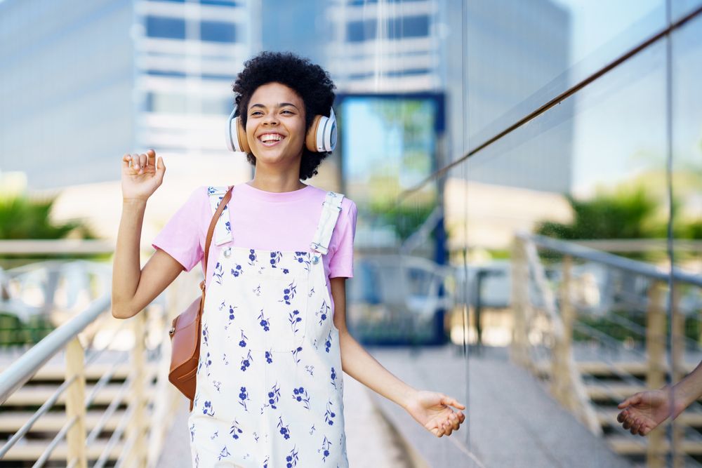Joyful female in floral overalls walking outside past corporate building listening to headphones