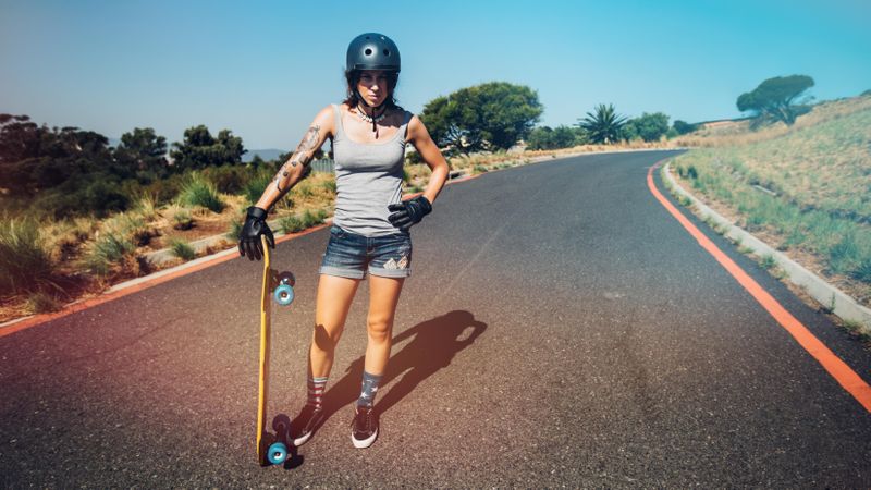 Full length portrait of young woman in helmet standing on a countryside highway with longboard