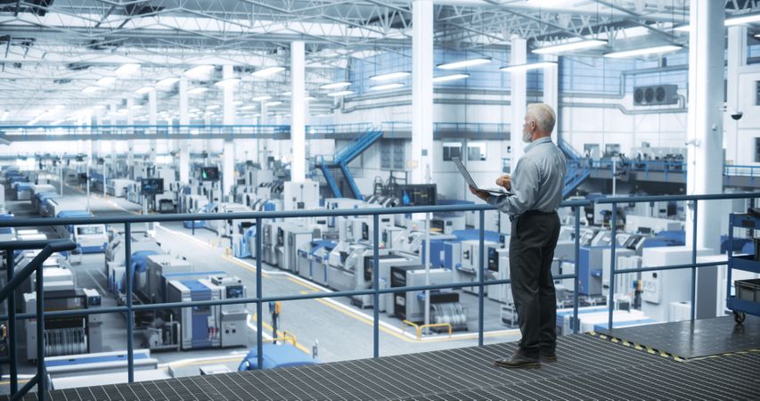Mature Engineer Standing on a Platform, Using Laptop Computer at an Electronics Factory. Machines are Undergoing Maintenance, Specialist Monitoring the Progress Through Online Software