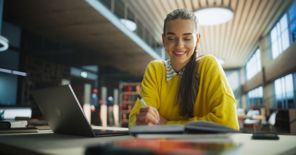 Happy and Focused Student Studying in a Modern Library. Young Female Enjoying Time Alone for Thinking and Problem Solving. Girl Using Laptop Computer to Work on a University Research Project Online