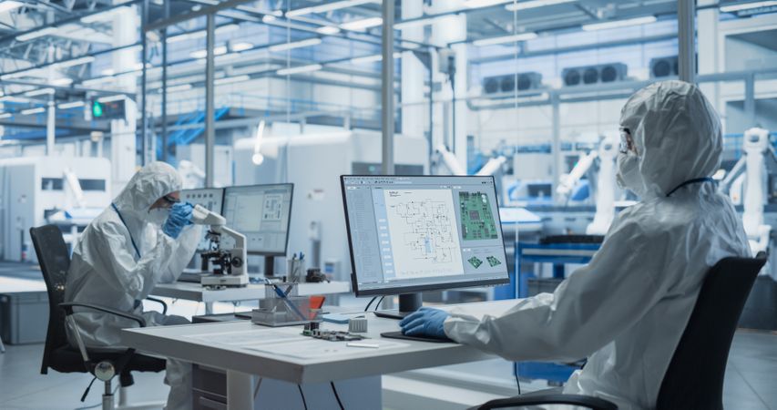 Establishing Shot of Two Laboratory Workers in Protective Coveralls Developing a New Advanced Central Processing Unit. PCB Design Engineer Working on a Computer, While Colleague is Using a Microscope