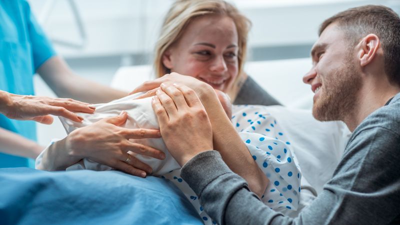 In the Hospital Midwife Gives Newborn Baby to a Mother to Hold, Supportive Father Lovingly Hugging Baby and Wife