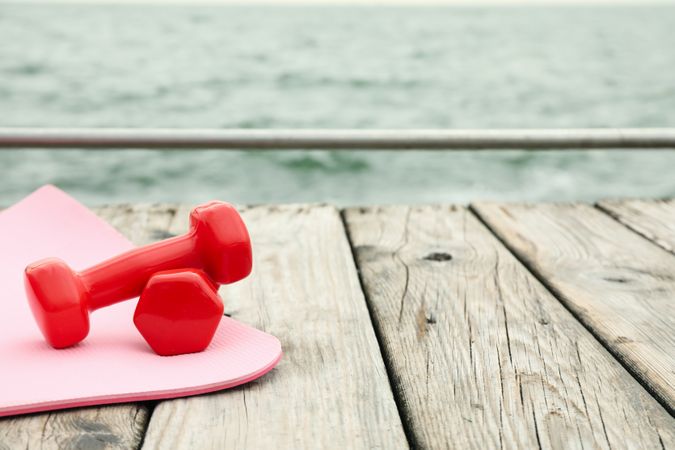 Yoga mat and dumbbells on wooden floor at sea, space for text