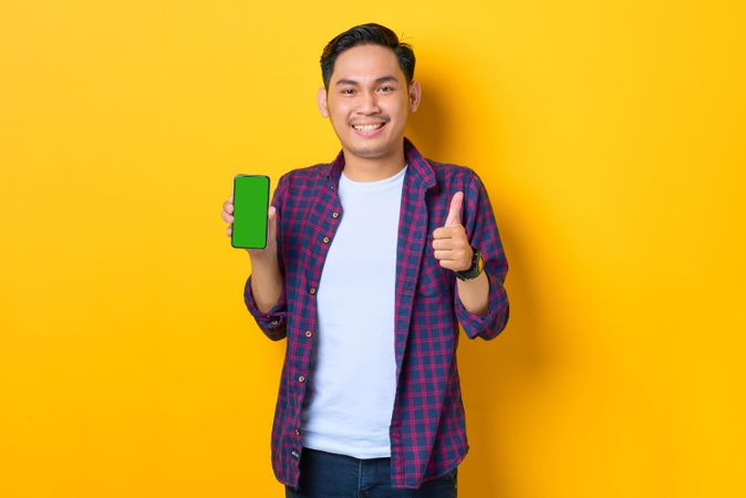 Asian man with thumbs up in plaid shirt holding smartphone with chroma key