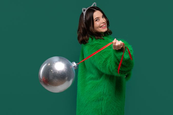 Woman in green coat holds silver Christmas ornament playfully