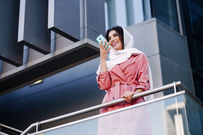 Female in headscarf and pink trench coat standing on staircase speaking on smartphone
