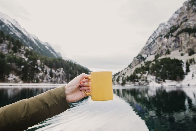 Person with cup near wonderful lake between hills in snow and cloudy sky