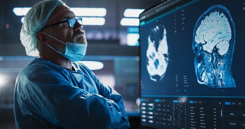 Male Neurosurgeon Looking At TV Screen With Brain MRI Scans Of Patient, Analyzing, Preparing For Complicated Neurosurgery, Wearing Surgical Gown.