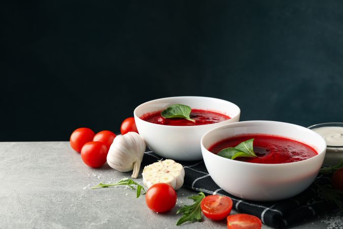 Tomato soup bowls surrounded by ingredients on grey kitchen counter, copy space