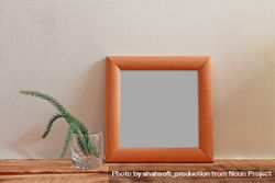 Plain square wooden picture frame with grey interior leaning against wall with plant in glass mockup 4NDpr0