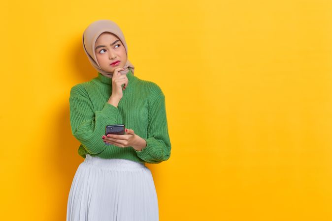 Woman in headscarf pondering something holding smart phone with finger on chin