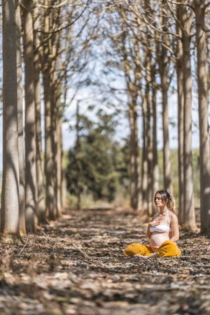 Calm adult pregnant woman practicing yoga while sitting in pose lotus on ground in park during sunny day