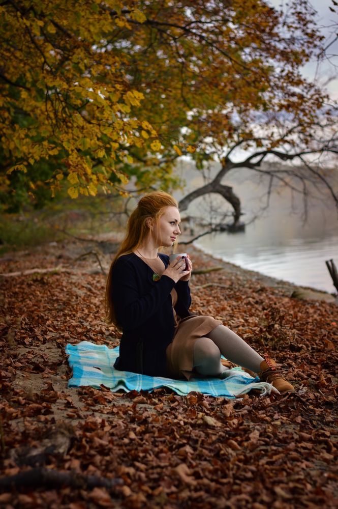 Woman holding a mug and sitting on autumn leaves beside waterside