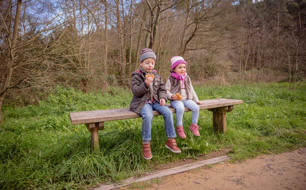 Young brother and sister sitting on park bench talking with snack