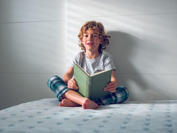 Cheerful curly blond boy reading book while sitting on bed