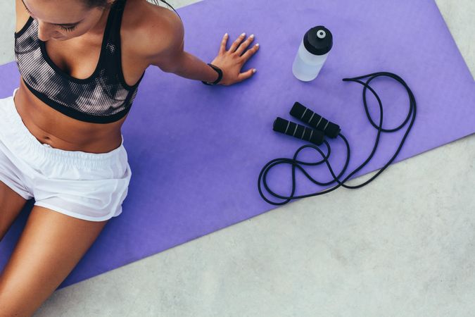 Top view of a fit woman sitting on a mat with water bottle and skipping rope beside her