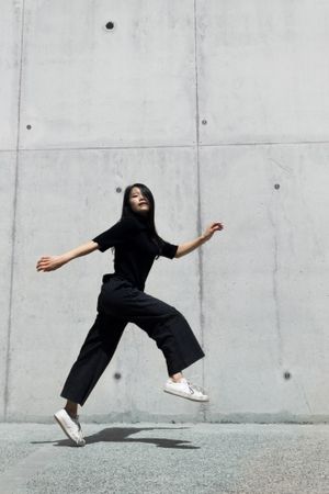 Woman in dark shirt and pants jumping outdoor near gray wall