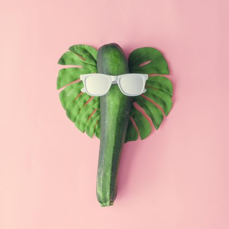 Monstera leaf, zucchini, and light painted sunglasses making face on pink background