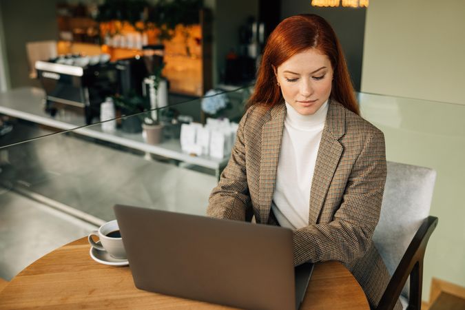 Woman in blazer working on laptop in cafe with coffee