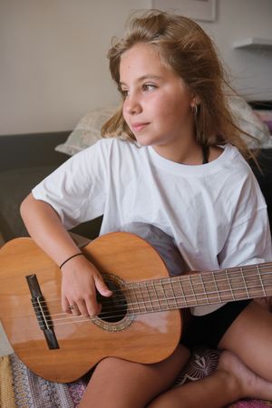 Vertical photo of a blonde girl looking to a side with distracted expression while playing a guitar