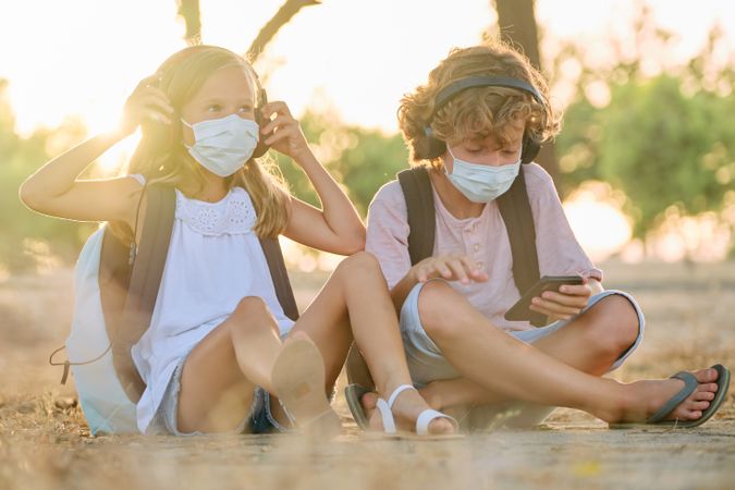 Brother and sister in masks sitting on ground in woods