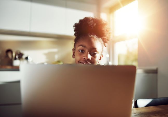 Shot of happy girl peaking over laptop in sunny kitchen