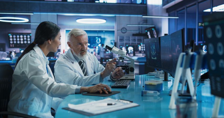 Biotechnology Medical Research Center: Female Asian Neuroscientist Using Desktop Computer And Talking To Male Surgeon With Microscope.