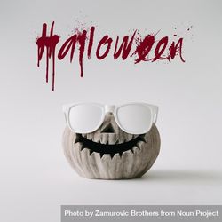 Pumpkin skull with sunglasses on light wall with “Halloween” text bGWLvb