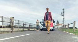 Male in red checkered shirt walking in front of broken down van with gas can