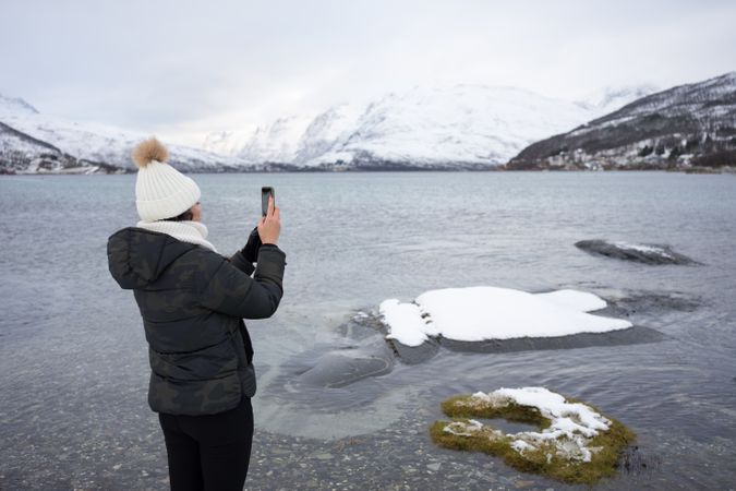 Woman shooting with smartphone on pond beach against snowy hills in cold overcast weather