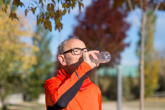 Grey haired man in red shirt and glasses sipping from water bottle in park
