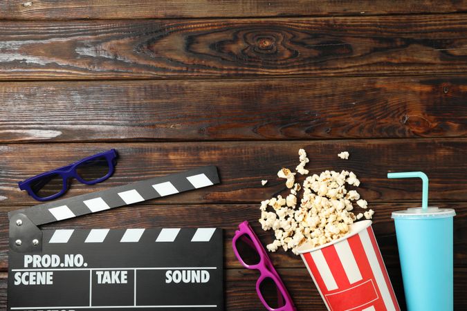 Flat lay. Bucket with popcorn, clapperboard, 3d glasses and drink on wood background