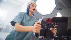 Focused Young Female Videographer With Blue Hair Adjusts Her Camera During An Indoor Film Shoot, Capturing The Essence Of Modern Filmmaking And Creative Production In A Professional Setting.