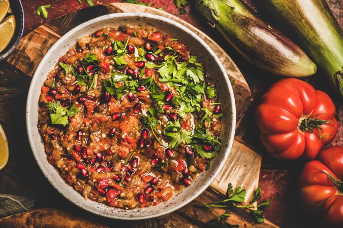 Eggplant dip with pomegranate and parsley garnish on wooden surface with vegetables