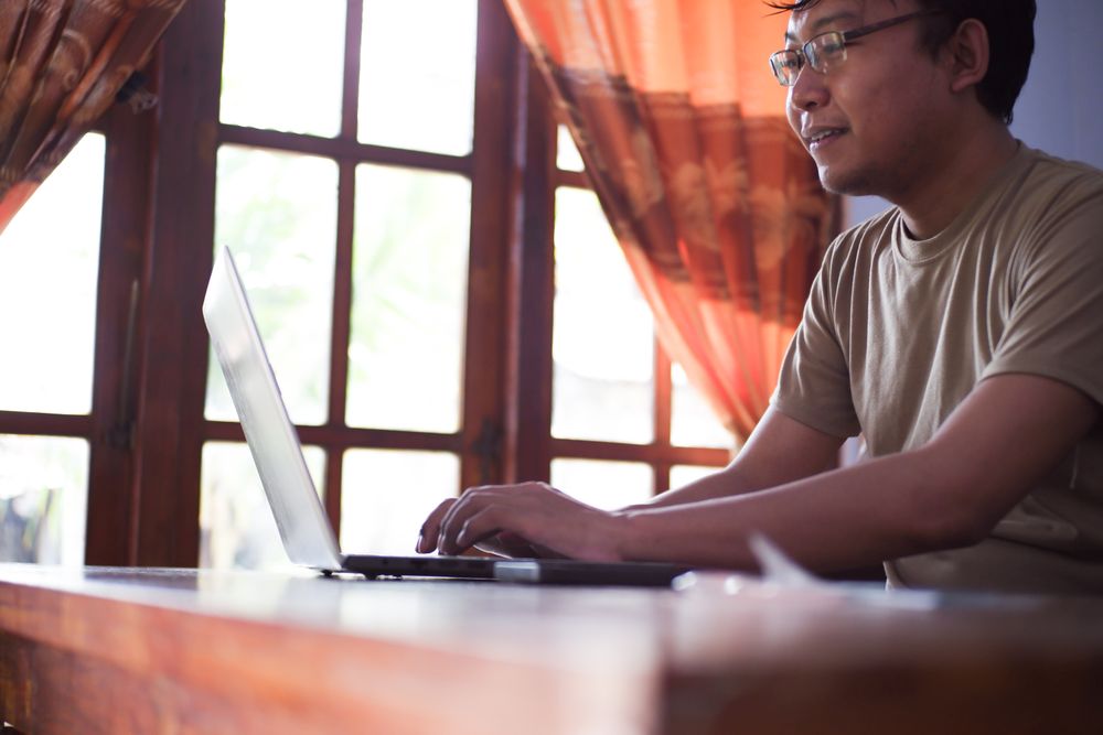 Asian Male with shirt working from home in living room with book, laptop and cup of coffee