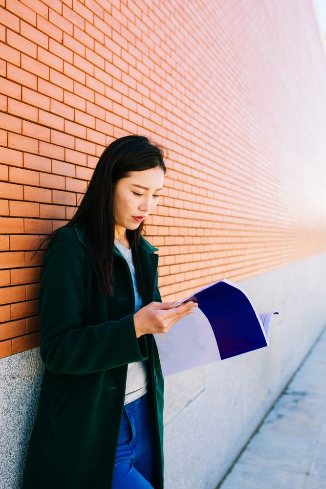 Asian student reading textbook near brick wall