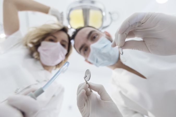 Dentists wearing surgical mask while holding angled mirror and drill, ready to begin
