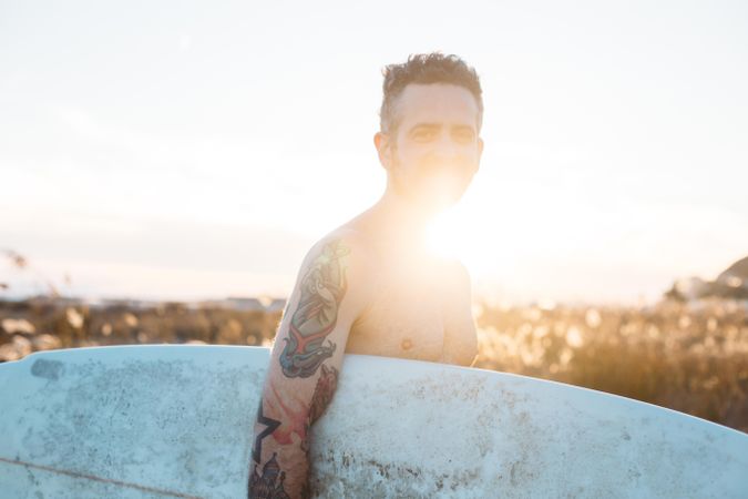 Tattooed man with surfboard under his arm with sun at his back