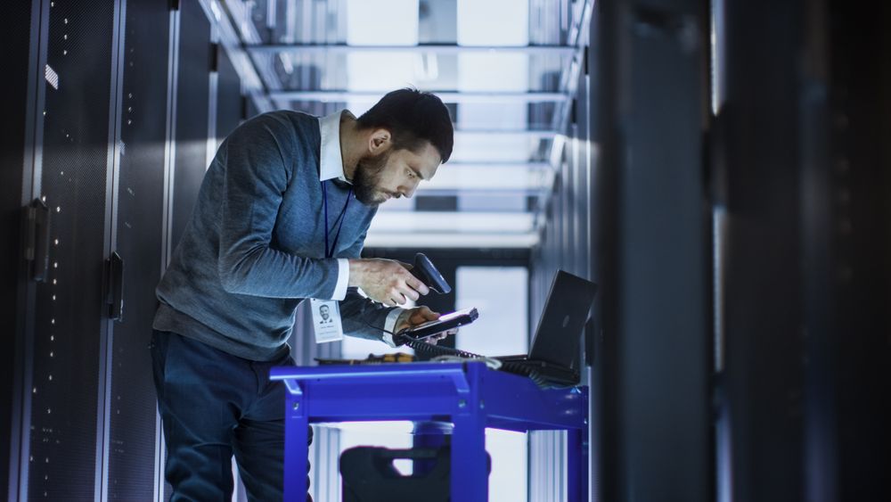 IT Engineer with Tool Cart Working on a Laptop Computer, he Holds a Hard Drive.