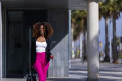 Black female in bright pink trousers with handbag standing outside on sunny day