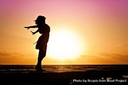 Silhouette of woman standing and opening her arms on beach during sunset 0PWxlb