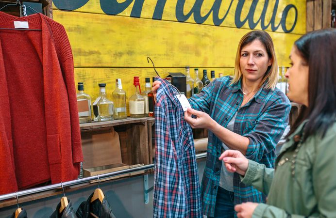 Woman shop assistant showing label price of plaid shirt to client in shop