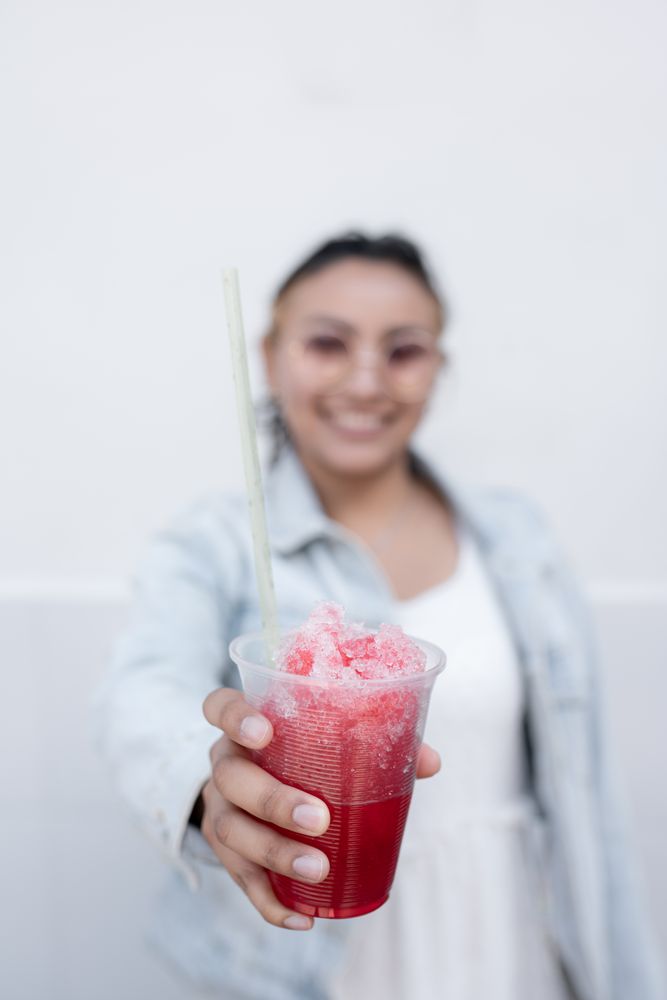 A young Hispanic woman is smiling while drinking a Mexican cherry slushy outdoors
