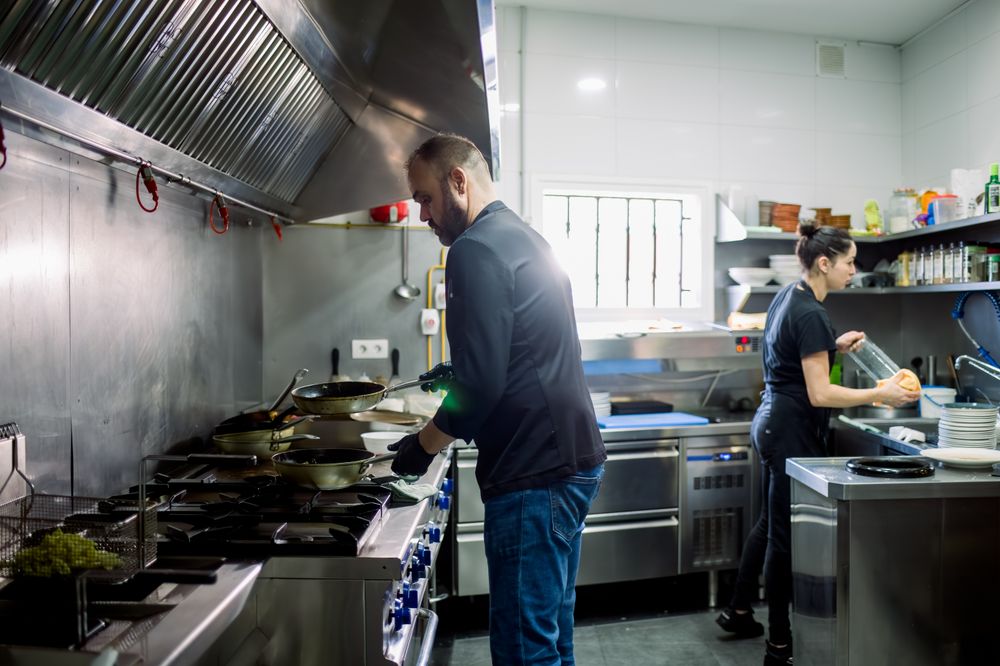 Side view of bearded male chef and woman cooking food over pan and washing dishes in modern kitchen of restaurant